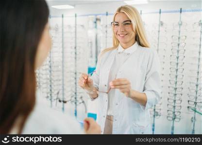Female optician and customer shooses glasses in optics store. Selection of eyeglasses with professional optometrist. Eye care, spectacles choice. Female optician and customer shooses glasses