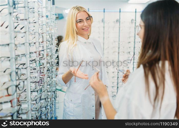 Female optician and customer shooses glasses in optics store. Selection of eyeglasses with professional optometrist. Eye care, spectacles choice