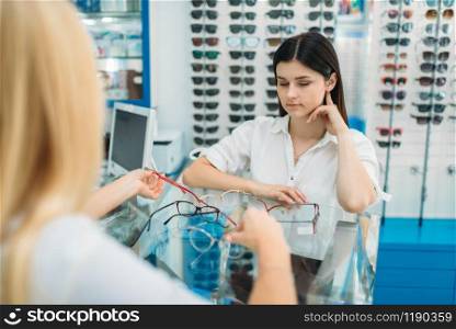 Female optician and woman chooses glasses frame in optics store. Selection of spectacles with professional optometrist