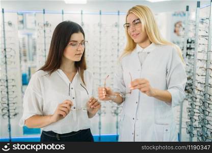 Female optician shows glasses to customer in optics store. Selection of eyeglasses with professional optometrist. Eye care