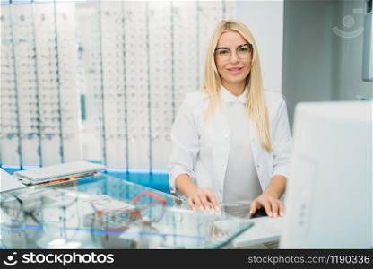 Female optician sitting at the table, showcase with spectacles in optics store. Selection of eyeglasses with professional optometrist