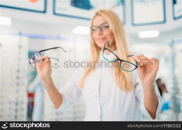 Female optician standing against showcase with glasses in optics store. Selection of eyeglasses with professional optometrist. Female optician against showcase with glasses