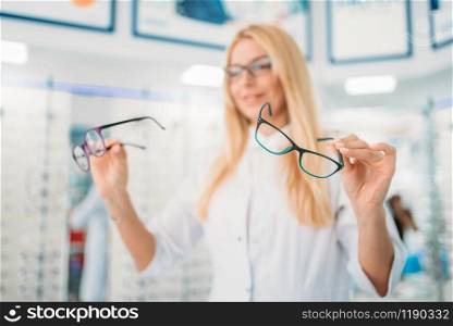 Female optician standing against showcase with glasses in optics store. Selection of eyeglasses with professional optometrist. Female optician against showcase with glasses