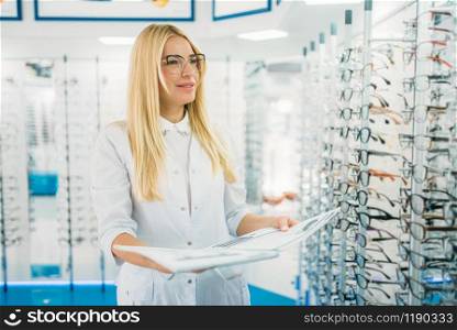 Female optician with glasses catalog in hands standing against showcase with spectacles in optics store. Selection of eyeglasses with professional optometrist