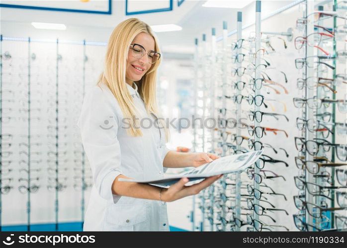 Female optician with glasses catalog in hands standing against showcase with spectacles in optics store. Selection of eyeglasses with professional optometrist. Female optician with glasses catalog in hands