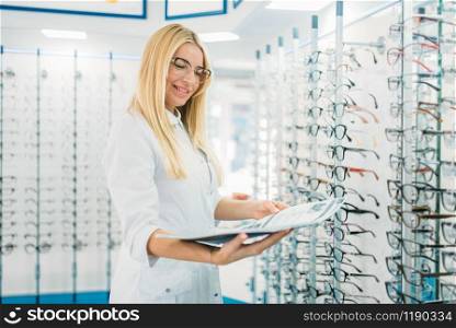 Female optician with glasses catalog in hands standing against showcase with spectacles in optics store. Selection of eyeglasses with professional optometrist. Female optician with glasses catalog in hands