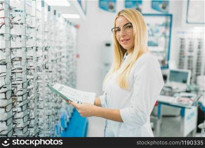Female optician with glasses catalog in hands standing against showcase with spectacles in optics store. Female optician with glasses catalog in hands