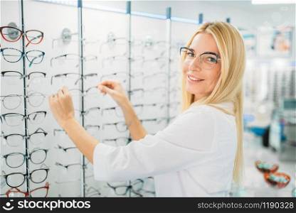 Female optometrist shows glasses in optics store. Selection of eyeglasses with professional optician, optometry