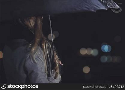 Female outdoors in rainy night, rear view of the elegant woman under black umbrella looking on the city lights, city life, loneliness and melancholy concept. Female outdoors in rainy night
