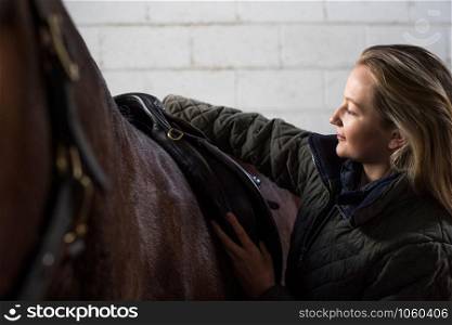 Female Owner Adjusting Saddle Straps In Stable With Horse