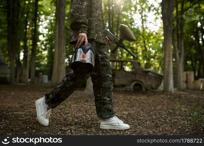 Female paintball player holds gun and mask, playground in the forest on background. Extreme sport with pneumatic weapon and paint bullets or markers, military team game outdoors, combat tactics. Female paintball player holds gun and mask