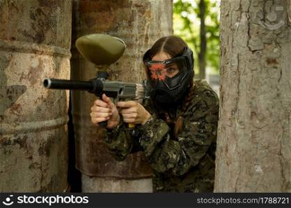 Female paintball player with gun poses on playground in the forest. Extreme sport with pneumatic weapon and paint bullets or markers, military team game outdoors, combat tactics. Female paintball player on playground in forest