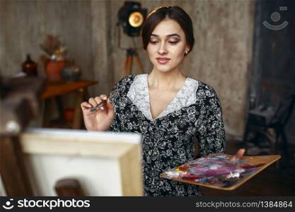Female painter with brush and palette draws in art studio. Artist at her workplace, creative master at the easel in workshop. Female painter with brush and palette in studio