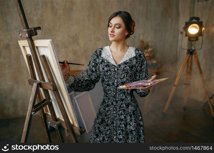 Female painter with brush and palette draws in art studio. Artist at her workplace, creative master at the easel in workshop. Female painter with brush and palette in studio