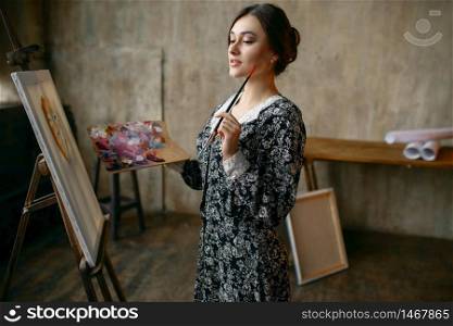 Female painter with brush and palette poses in art studio. Artist at her workplace, creative master at the easel in workshop. Female painter poses in art studio