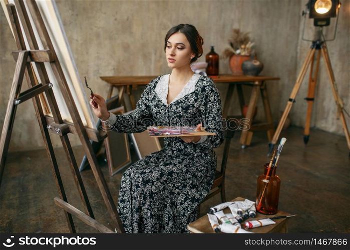 Female painter with brush and palette poses in art studio. Artist at her workplace, creative master at the easel in workshop. Female painter poses in art studio