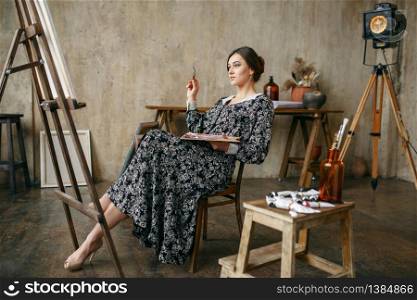 Female painter with brush and palette sitting at the easel in art studio. Artist at her workplace, creative master in workshop. Female painter sitting at the easel in art studio