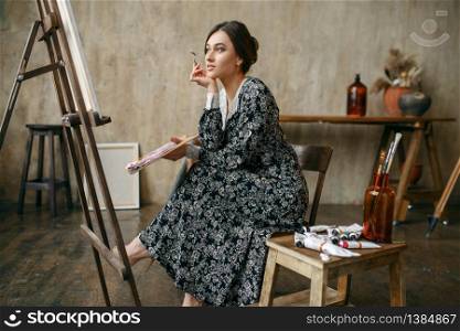 Female painter with brush and palette sitting at the easel in art studio. Artist at her workplace, creative master in workshop. Female painter sitting at the easel in art studio