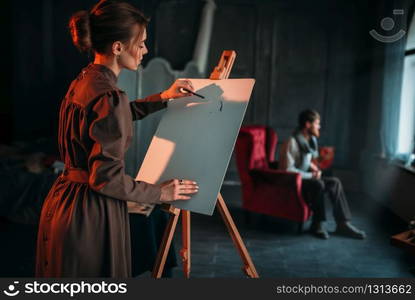 Female painter with palette and brush in hand paints man portrait on easel in art studio. Oil paint, paintbrush drawing. Female painter paints man portrait in art studio