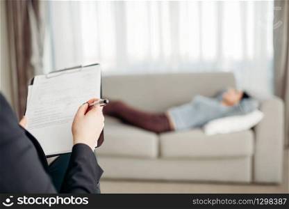 Female patient lies on sofa at psychotherapist reception. Female doctor writes notes in notepad, professional psychology support. Patient lies on sofa at psychotherapist reception