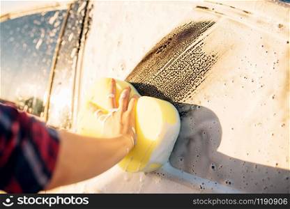 Female person hand with sponge scrubbing vehicle with foam, car wash. Young woman on self-service automobile washing. Outdoor carwash. Female person scrubbing vehicle with foam