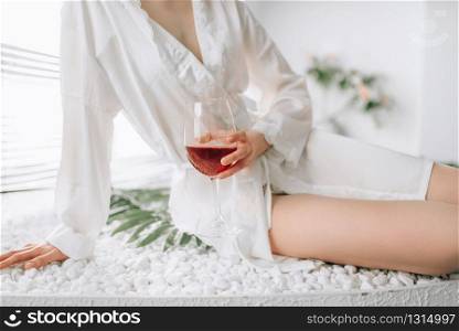 Female person in white bathrobe sitting on the edge of the bath with glass of red wine. Bathroom interior with window on background