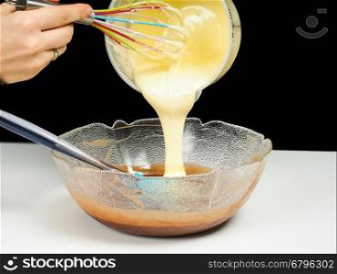 Female person pouring Gogl-Mogl, into a glass bowl with chocolate, with hand whisker from plastic bowl, isolated on black