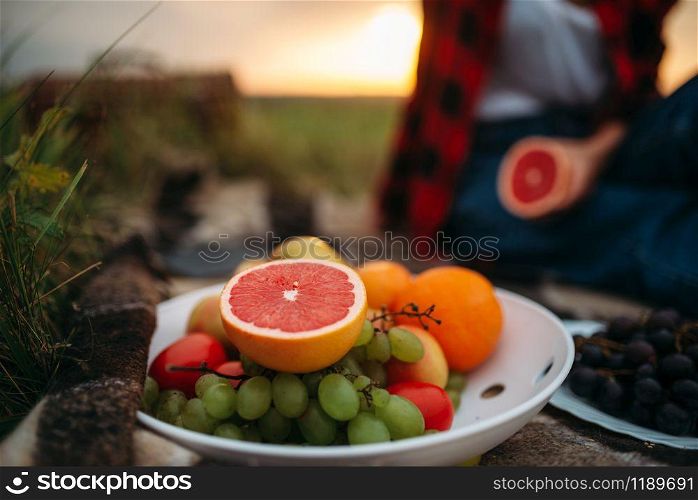 Female person with fruits sitting on plaid, picnic in summer field. Romantic junket, happy holiday