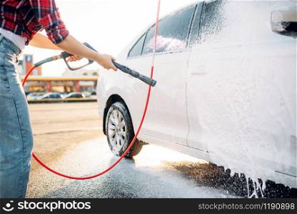 Female person with high pressure water gun in hands wash off the foam from the car. Young woman on self-service automobile washing. Outdoor vehicle cleaning at summer day