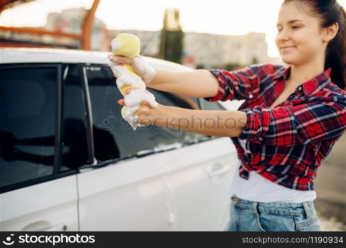 Female person with sponge scrubbing vehicle window with foam, car wash. Young woman on self-service automobile washing. Outdoor carwash. Female person scrubbing vehicle window with foam