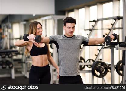 Female personal trainer helping a young man lift dumbells while working out in a gym