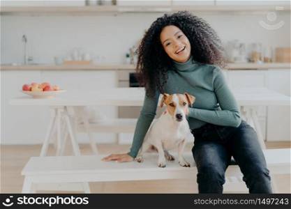 Female pet owner expresses love to dog, dressed in casual turtleneck and jeans, sits at white bench against cozy kitchen interior, spends free time at home, smiles broadly from happy enotions