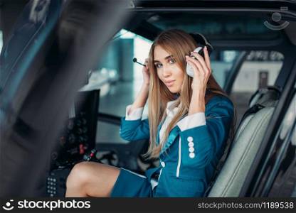 Female pilot in headphones poses in helicopter cabin, hangar interior on background, view from cabin. Air hostess in uniform sits in copter. Private airline. Female pilot in headphones in helicopter cabin