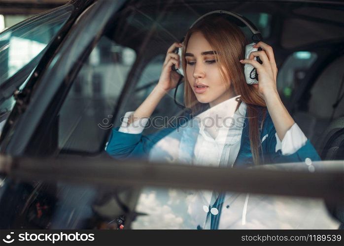 Female pilot in headphones sits in helicopter. Air hostess in uniform in copter. Private airline. Female pilot in helicopter, view from windshield