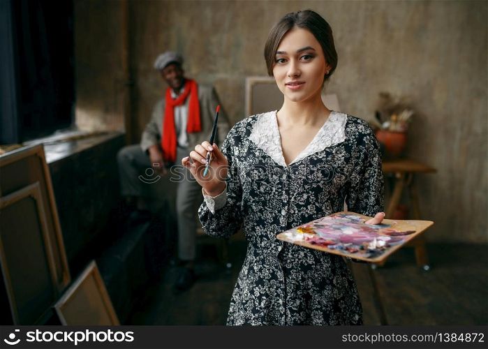 Female portrait painter holds palette and brush, male model in art studio on background. Male artist standing at his workplace, creative master in workshop. Female portrait painter holds palette and brush