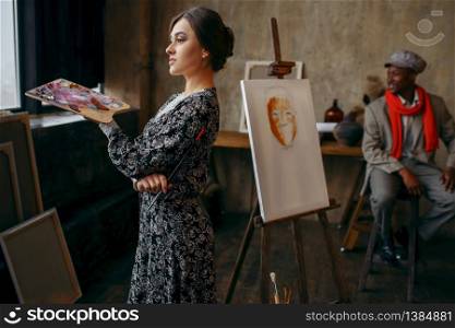 Female portrait painter holds palette and brush, male model in art studio on background. Male artist standing at his workplace, creative master in workshop. Female portrait painter holds palette and brush