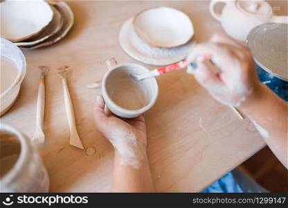 Female potter with brush paints a pot, pottery workshop. Woman molding a bowl. Handmade ceramic art, tableware from clay. Female potter with brush paints a pot, pottery