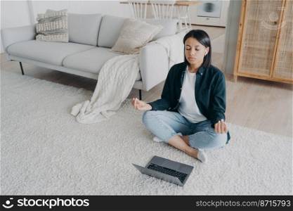 Female practices yoga at laptop on the floor, listening meditation to relieve stress after work at home. Calm woman in earphones meditates sitting in lotus pose in front of computer. Healthy lifestyle. Female practices yoga at laptop on floor, listening meditation to relieve stress after work at home