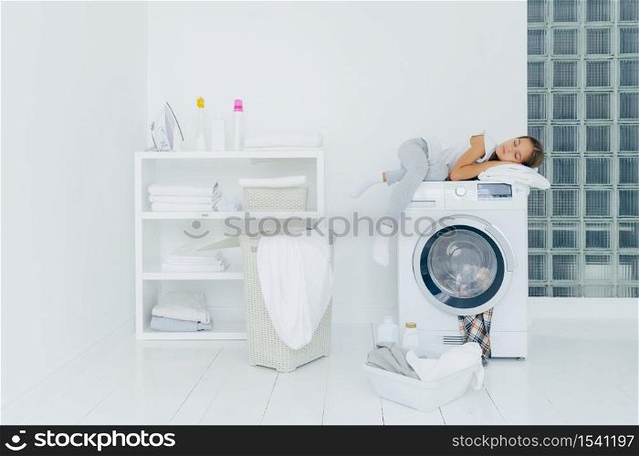 Female preschooler sleeps on washing machine, being tired with washing, poses in white big laundry room with basket and basin full of dirty clothes bottles of liquid powder. Childhood, domestic chores