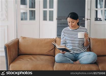 Female reads bestseller book relaxing sitting on couch at home. Pensive woman enjoying reading novel, resting with coffee cup on comfortable sofa in living room on weekend. Hobby, education concept.. Woman rests reading bestseller novel holding paper book, coffee cup, sitting on sofa at home