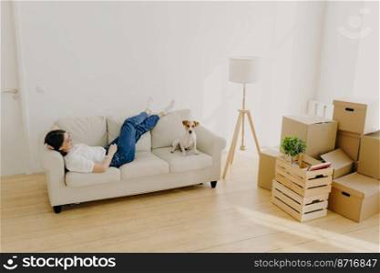 Female renter lies on white comfortable couch, raises crossed legs, poses with favourite dog, has new dwelling in living room, enjoys relocation day, feels relaxed. New home and relocation concept