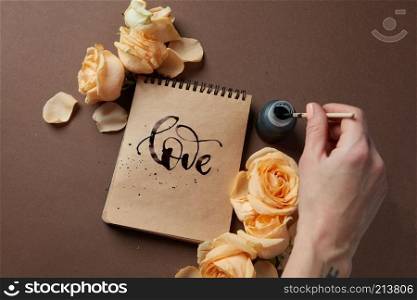 Female&rsquo;s hand using ink for writing word love in diary or notebook in Valentine&rsquo;s Day. Notebook with brown pages represented with orange flowers.. Diary or notebook with word love