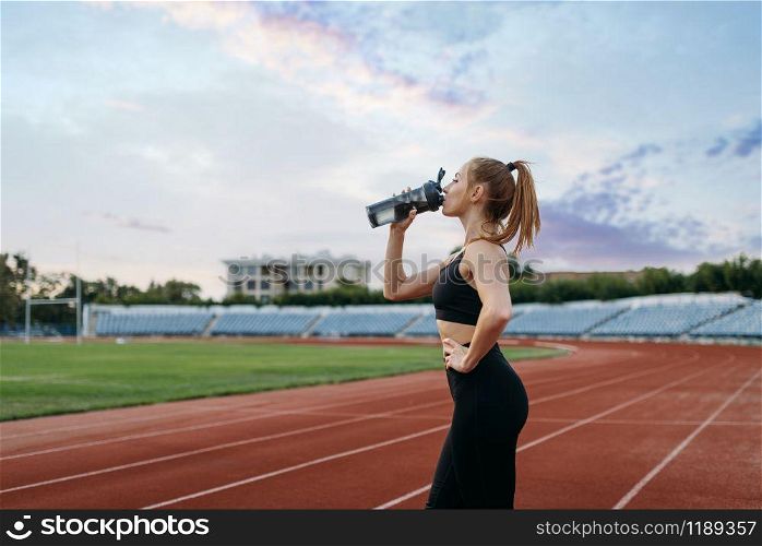 Female runner in sportswear drinks water, training on stadium. Woman doing stretching exercise before running on outdoor arena. Female runner drinks water, training on stadium