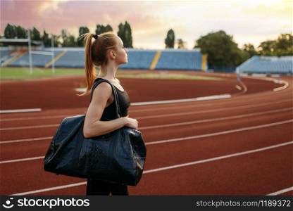 Female runner in sportswear holds sport bag, back view, training on stadium. Woman doing stretching exercise before running on outdoor arena. Female runner holds sport bag, training on stadium