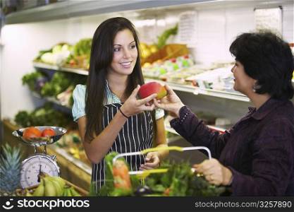 Female sales clerk giving a mango to a mature woman in a grocery store