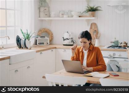 Female sales manager is working from home. Young spanish woman in orange shirt is remote worker. Lady freelancer is taking notes and browsing internet using laptop.. Female sales manager is working from home. Lady freelancer with laptop is taking notes.