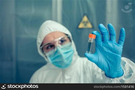 Female scientist with bacteriological protection suit watching vial in the laboratory. Selective focus on vial in foreground