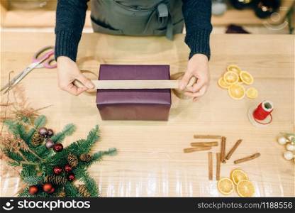 Female seller decorates gift box with wrapping paper and golden ribbon, decoration process. Woman wraps present on the table, decor procedure. Seller decorates gift box with golden ribbon