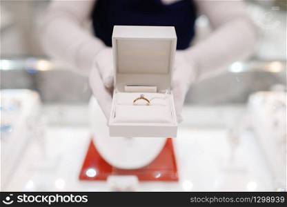 Female seller hands holds gold diamond ring in a box, jewelry store. Salewoman at the showcase holds decorative case with golden decoration, jewellery shop