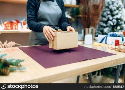 Female seller holds unwrapped carton gift box. Present wrapping papaer on the table, decoration procedure